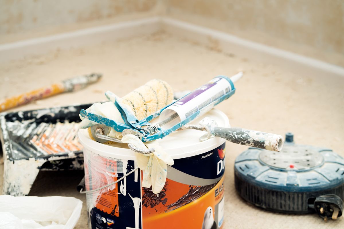 Paint bucket with a roller sleeve, caulk gun, scraper, and gloves resting on top, and an extension cord reel nearby